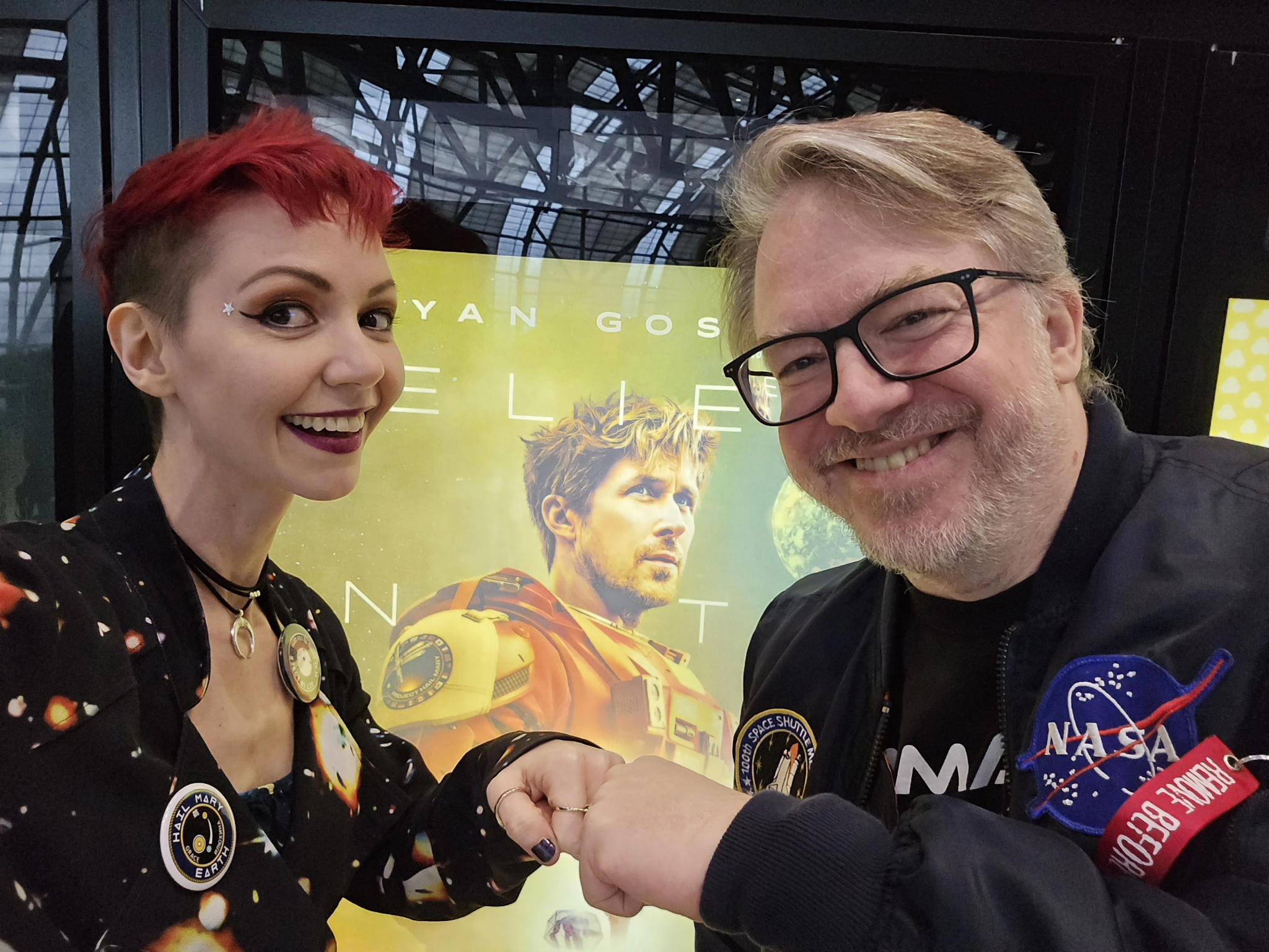 Jennie with short red hair and a space-themed jacket with a Project Hail Mary pin and James in a NASA bomber jacket excitedly fist bump in front of the Project Hail Mary poster at the cinema.