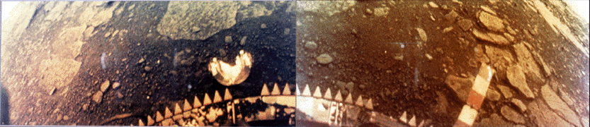 A colour 170 degree panorama displaying brown and red flat rock slabs and soil. The top of a portion of the spacecraft with many small triangular protrusions is visible at the bottom of the image. A round camera lens cap is visible on the soil, amid rocks and pebbles.