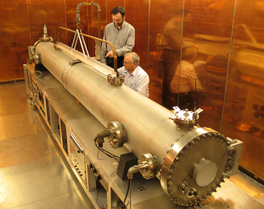 Two scientists wearing long-sleeved button-up shirts inspect the atomic clock, seen as a long, golden, cylindrical vacuum chamber atop a reflective metallic surface. The room itself, including the walls and ceiling, also appears metallic.