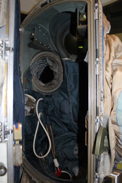 The backpack hatch of a white Orlan suit hangs open, revealing the navy blue and grey internal compartment. Inside the hatch are a few metal coiled wires, a white looped tube, and some other cables that appear to be insulated in blue fabric. Holes for the left arm and head lead to dark compartments. On the left side, the edge of the white backpack door is edge-on to the camera. On the right of the image is a portion of the white external right arm of the suit.