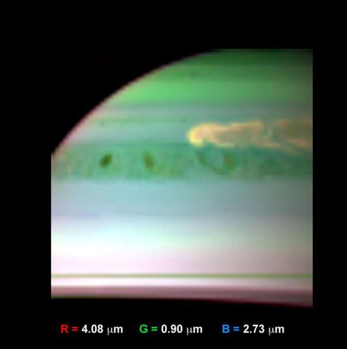 Approximately one-quarter of the surface of Saturn, appearing to be the upper-left quadrant from the camera's perspective, is shown. The top of Saturn is largely green with a gradient that fades down through blue and yellow and pink. Toward the very bottom of the image, sharp horizontal lines are shown, presumably the rings seen nearly-edge on. A little over halfway up the image is a diffuse band of greenish-yellow blobs in the atmosphere, beneath a swirling yellow cloud shape. Below the image itself, a guide to the false colors is given, with red representing 4.08 micrometers, green representing 0.90 micrometers, and blue representing 2.73 micrometers.