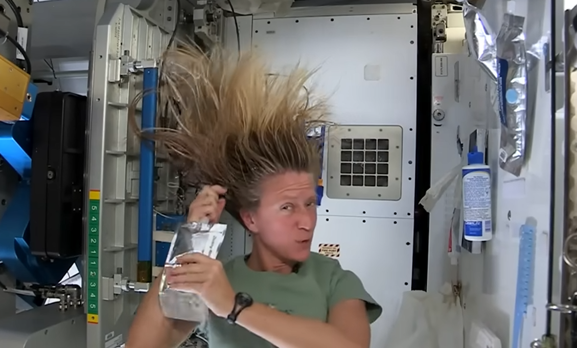 Astronaut Karen Nyberg is featured from shoulders-up on the International Space Station. She is wearing an olive green t-shirt and a black wristwatch. Her slightly damp, long, blonde hair floats above her head as she squeezes water out of a metallic silver bag directly onto her scalp. Various pieces of equipment are affixed to the white and silver compartment surrounding her. A white bottle of no-rinse shampoo with a blue cap and blue accents is attached via velcro to the wall near her head.
