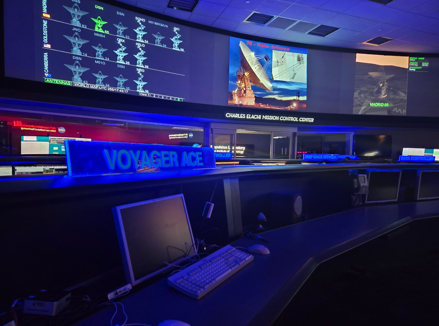Nearest to the observer is an elongated, curving desk containing computer monitors and one keyboard+mouse setup. The sign above the workstation reads 'VOYAGER ACE'. The entire room is coloured a cool shade of blue. On large curving screens above head-level against the far wall are displays related to the Deep Space Network. The leftmost screen shows the status of all antennas in Madrid, Goldstone, and Canberra. Each dish is coloured grey or white, though DSN 65 in Madrid appears in bright green. The middle screen displays an evening photograph of a large radio dish pointing up and to the right, with the red text '70-meter Antenna' just above it. Finally, the rightmost screen shows a visual of MADRID 65, the dish in dark grey shades and the text in bright green. Below the middle screen is a sign that reads 'CHARLES ELACHI MISSION CONTROL CENTER'.