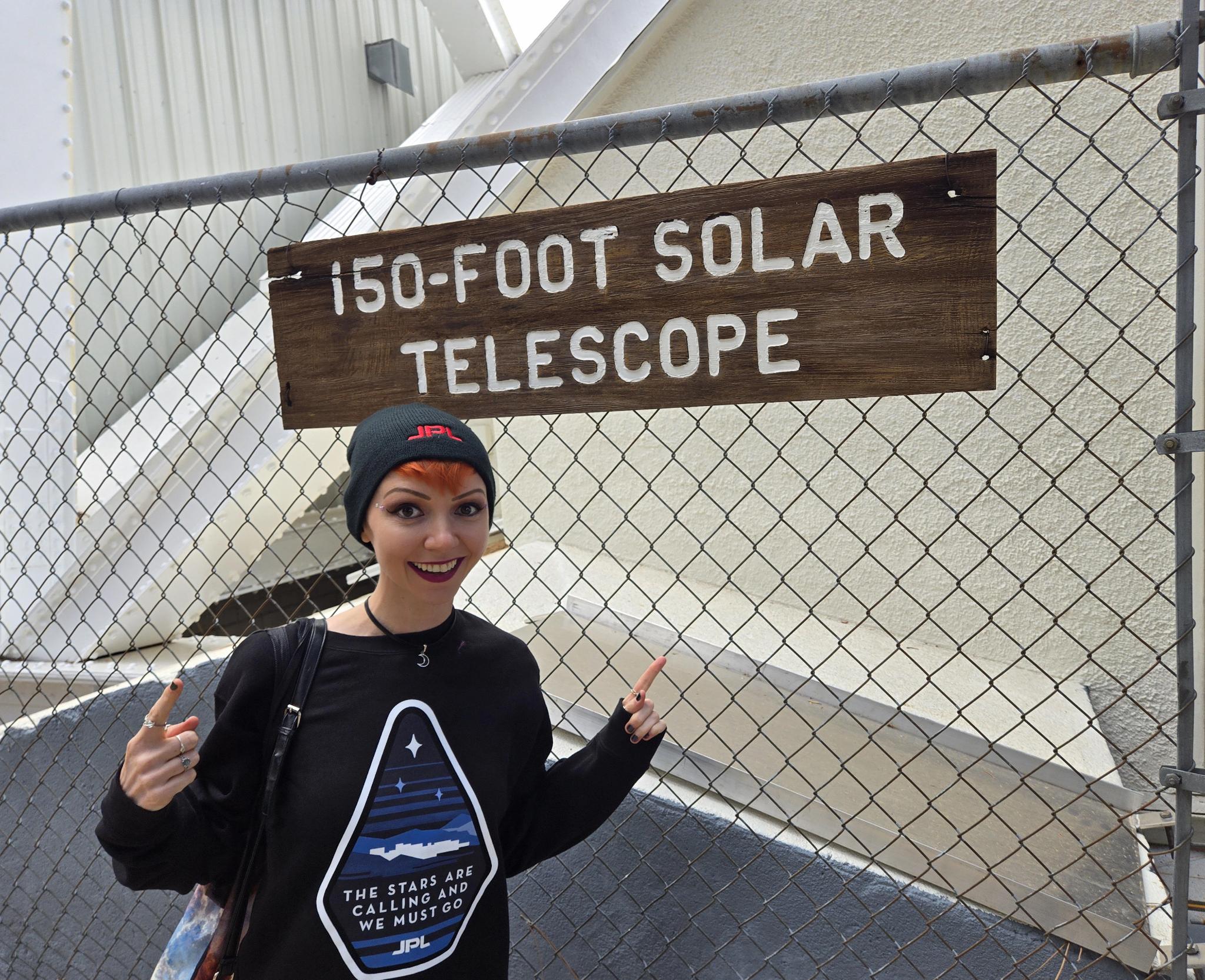 Jennie Starstuff, wearing a black JPL hat and a black JPL pullover that reads 'The stars are calling and we must go' points excitedly in front of a chain-link fence with an attached wooden sign that reads '150-foot solar telescope'. The white base of the telescope is visible behind the chain-link fence.