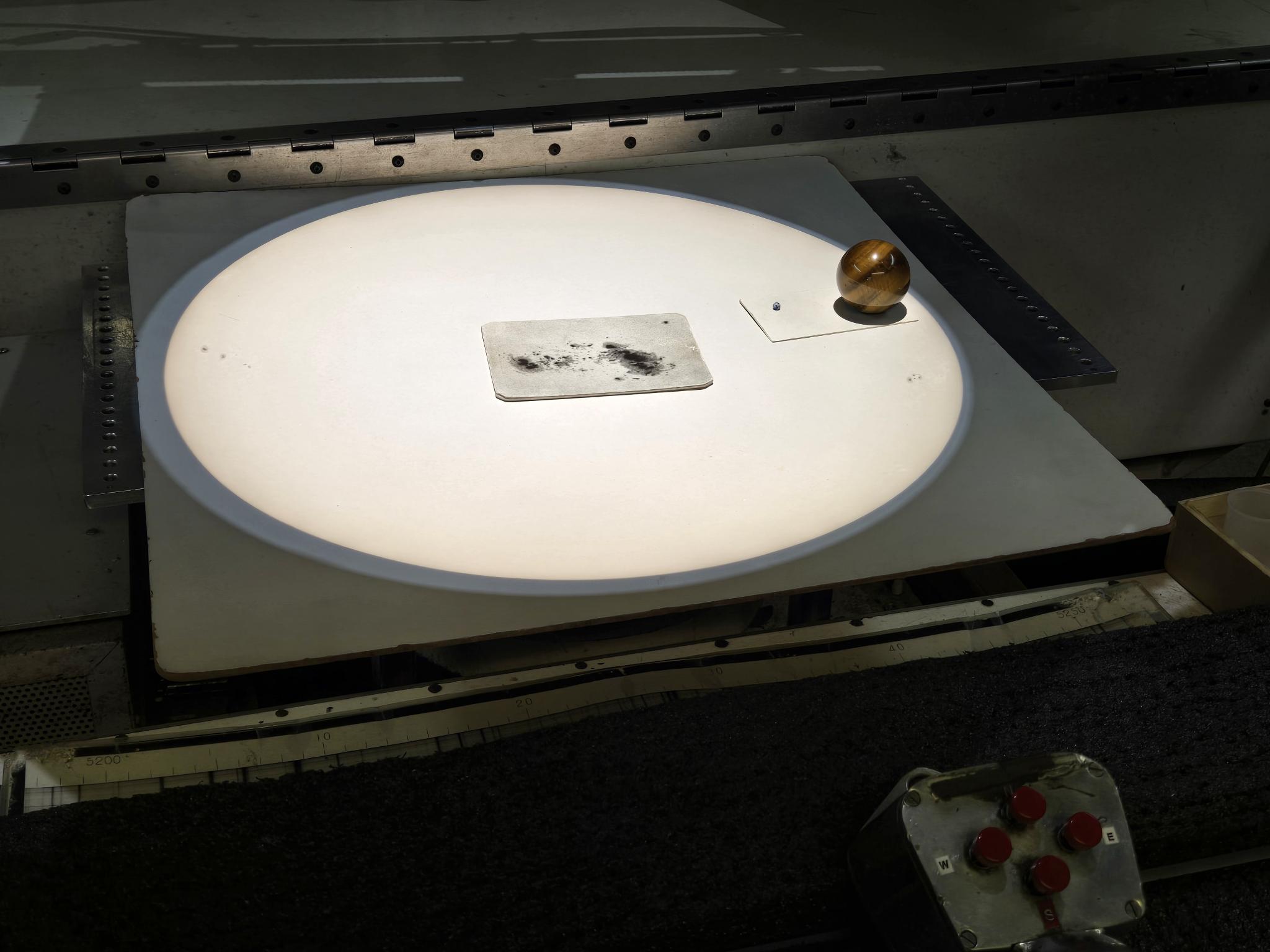 A bright white disc of the Sun is projected on a flat surface, inside the 150-foot solar telescope. Small sunspots can be seen on the Sun's left and right edges. In the center of the image is a card displaying a large irregularly shaped dark sunspot. To the right of the image are marbles representing the Earth and Jupiter.